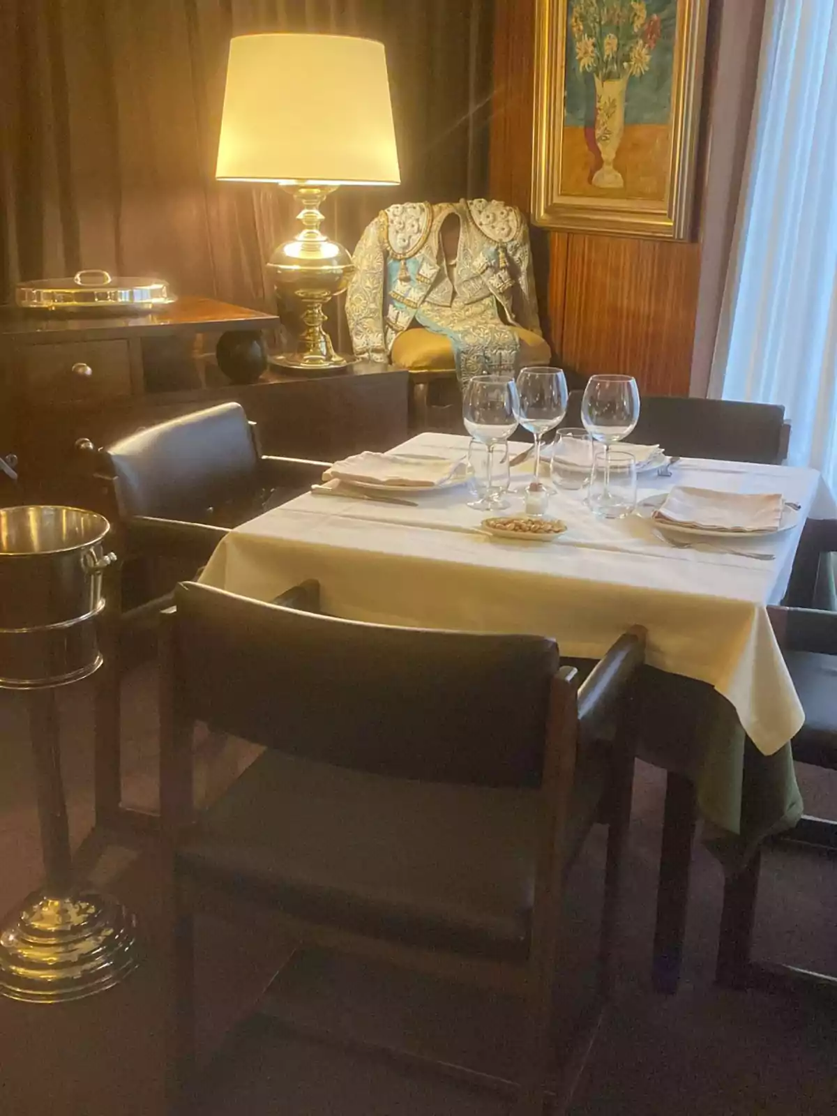 Una mesa de comedor con copas y platos, rodeada de sillas, con una lámpara encendida sobre un mueble, un traje de torero y un cuadro de flores en la pared.