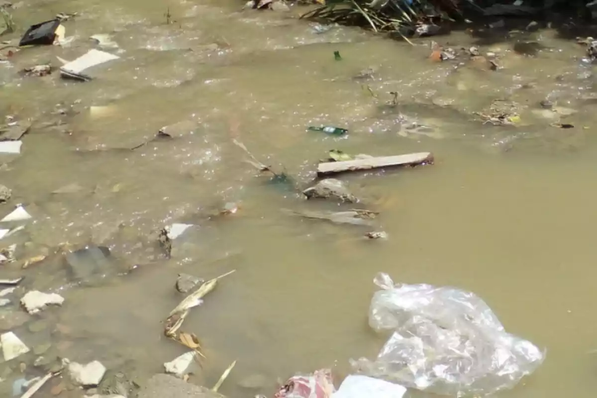 La imagen muestra un cuerpo de agua contaminado con diversos tipos de basura, incluyendo plásticos y escombros.