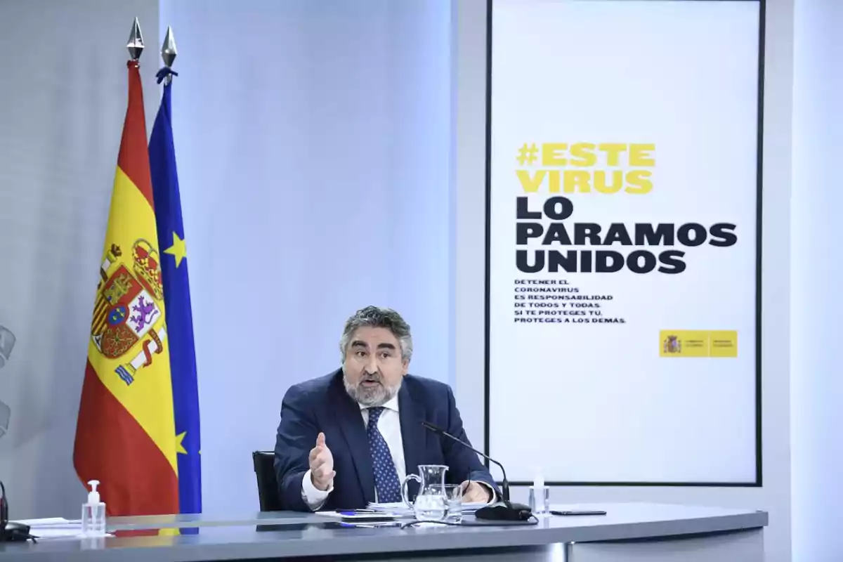 Un hombre en una conferencia de prensa con banderas de España y la Unión Europea detrás, junto a un cartel que dice 