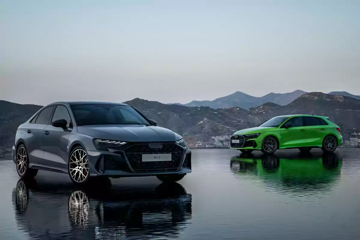 Dos autos Audi RS 3, uno gris y otro verde, estacionados sobre una superficie reflectante con montañas al fondo.