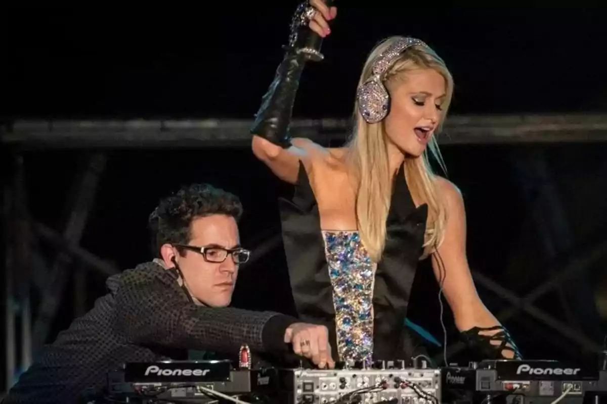 Una mujer rubia con auriculares brillantes está actuando como DJ en un escenario junto a un hombre que ajusta el equipo de sonido. Una mujer rubia con auriculares brillantes está actuando como DJ en un escenario junto a un hombre que ajusta el equipo de sonido.