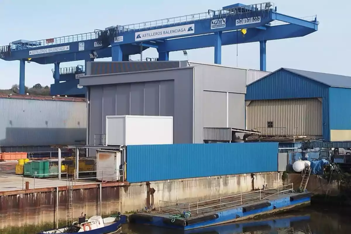 Una grúa azul de gran tamaño en un astillero junto a varios edificios industriales y una pequeña embarcación en el agua.