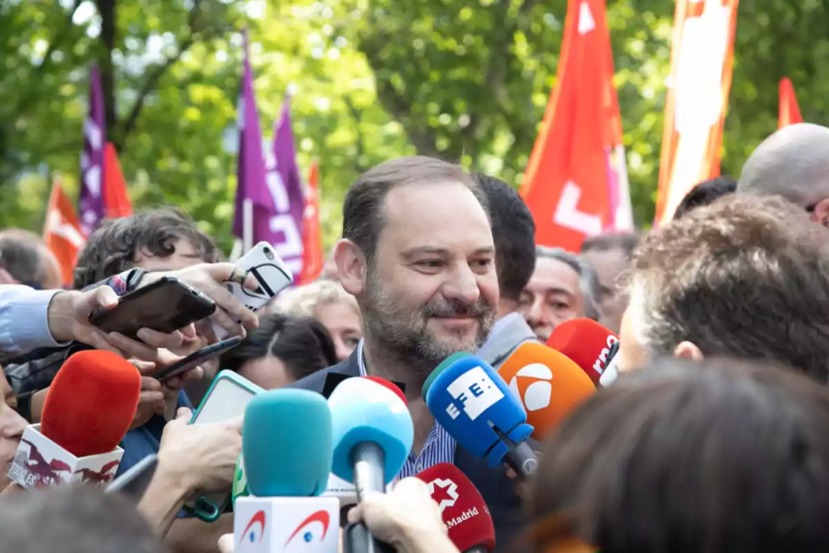 Un hombre sonriente rodeado de periodistas y micrófonos de diferentes medios de comunicación, con banderas de colores en el fondo.