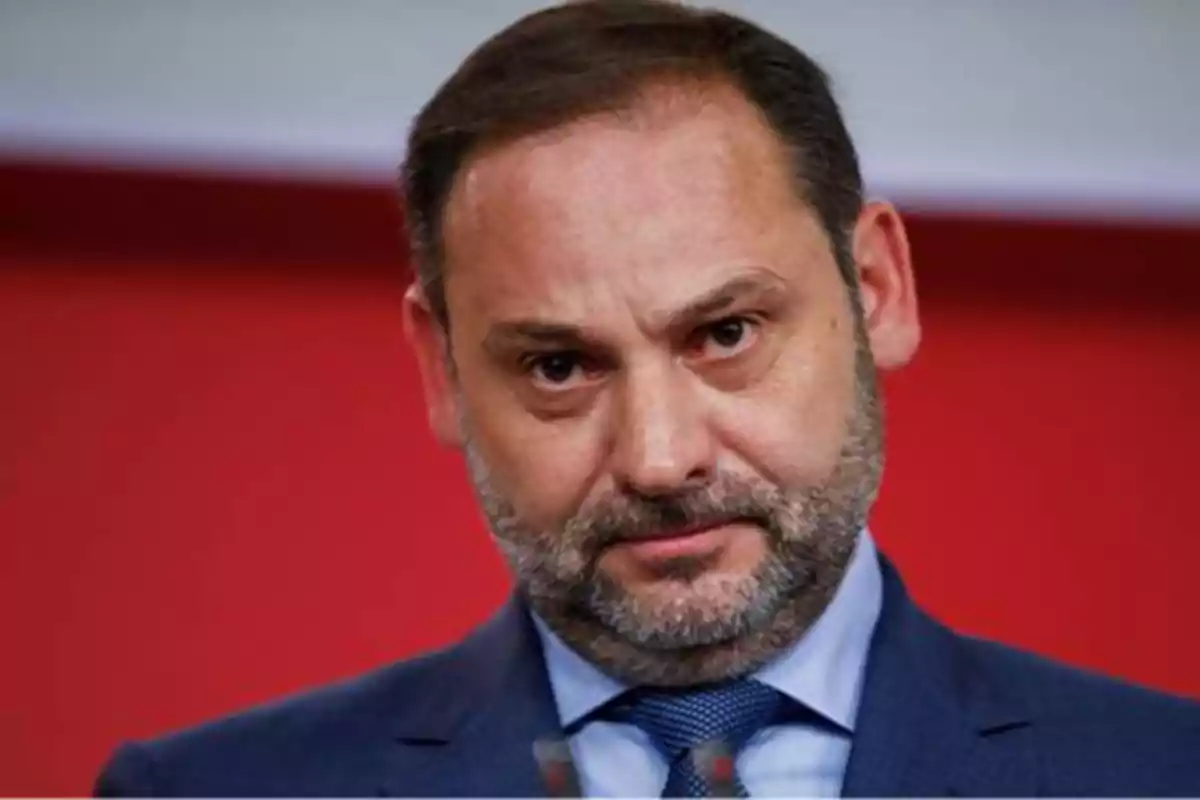 Un hombre con barba y traje azul mira hacia la cámara con expresión seria, sobre un fondo rojo.