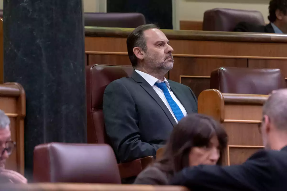 Un hombre con traje y corbata azul está sentado en un asiento de un recinto parlamentario.