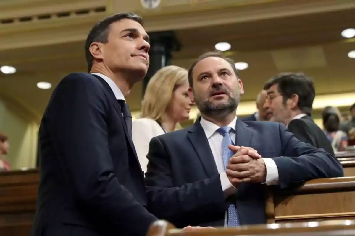 Dos hombres en traje conversan en un entorno formal con otras personas al fondo.