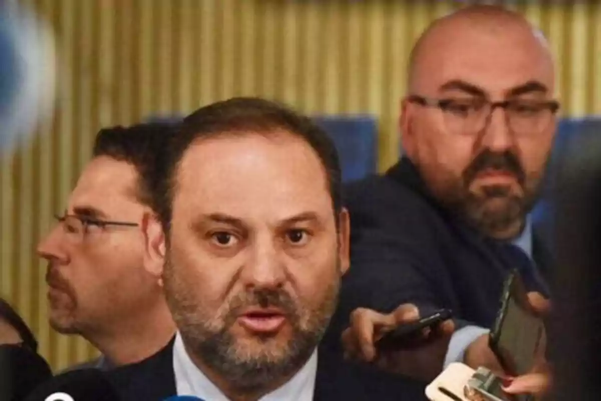 Un hombre hablando frente a varios micrófonos con personas al fondo.