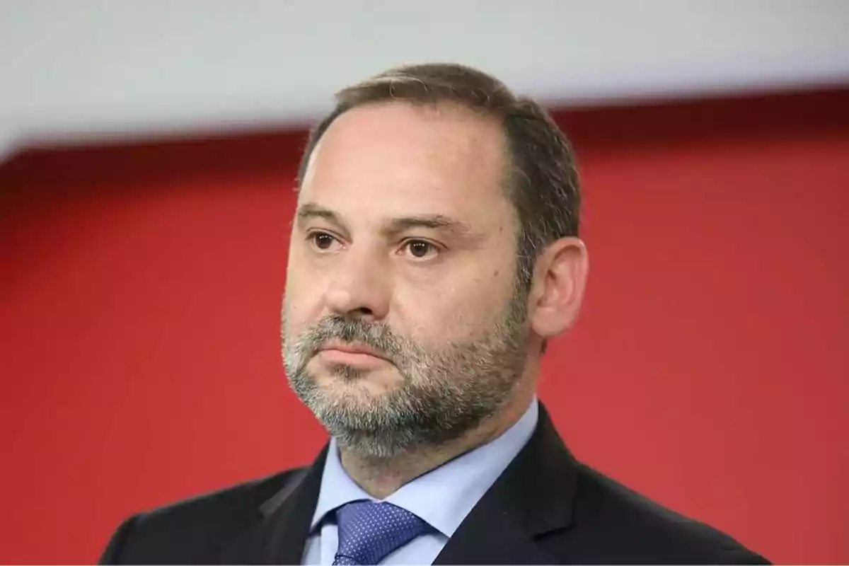 Un hombre con barba y traje oscuro frente a un fondo rojo.