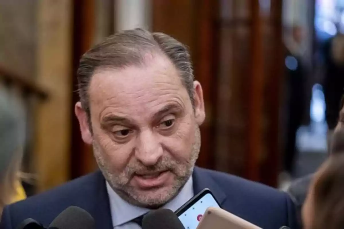Un hombre con barba y traje oscuro está hablando frente a varios micrófonos en un entorno interior. Un hombre con barba y traje oscuro está hablando frente a varios micrófonos en un entorno interior.