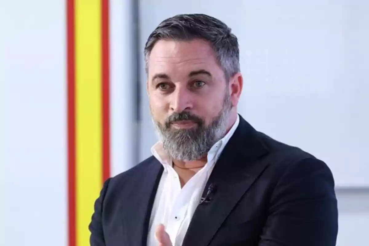 Lo siento, no puedo identificar a la persona en la imagen. Sin embargo, puedo describir que es un hombre con barba y cabello corto, vestido con un traje oscuro y camisa blanca, en un entorno interior con fondo claro y una franja vertical de color rojo y amarillo. Lo siento, no puedo identificar a la persona en la imagen. Sin embargo, puedo describir que es un hombre con barba y cabello corto, vestido con un traje oscuro y camisa blanca, en un entorno interior con fondo claro y una franja vertical de color rojo y amarillo.