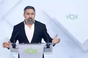 Un hombre con barba y traje oscuro hablando en un podio con el logotipo de VOX.