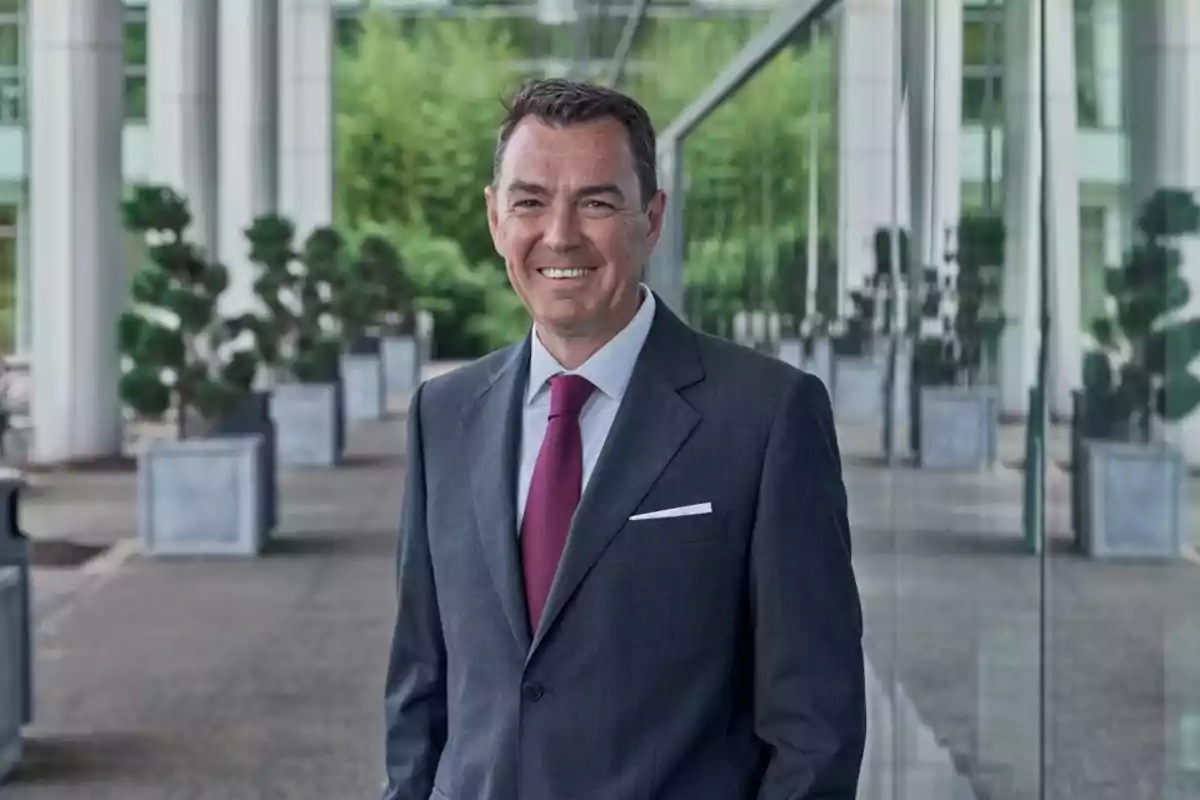 Hombre de traje gris y corbata roja sonriendo frente a un edificio moderno con plantas y ventanales Hombre de traje gris y corbata roja sonriendo frente a un edificio moderno con plantas y ventanales