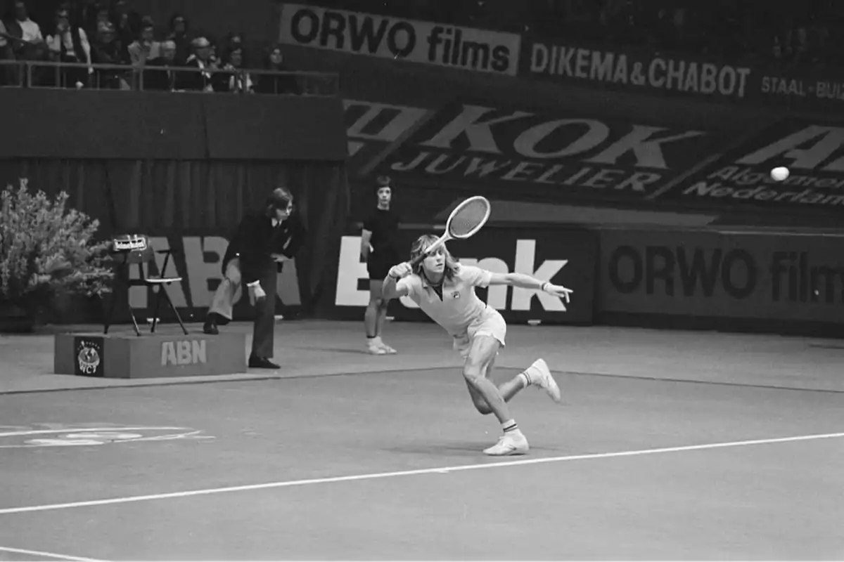 Un jugador de tenis se prepara para golpear la pelota en una cancha cubierta mientras el público observa desde las gradas.