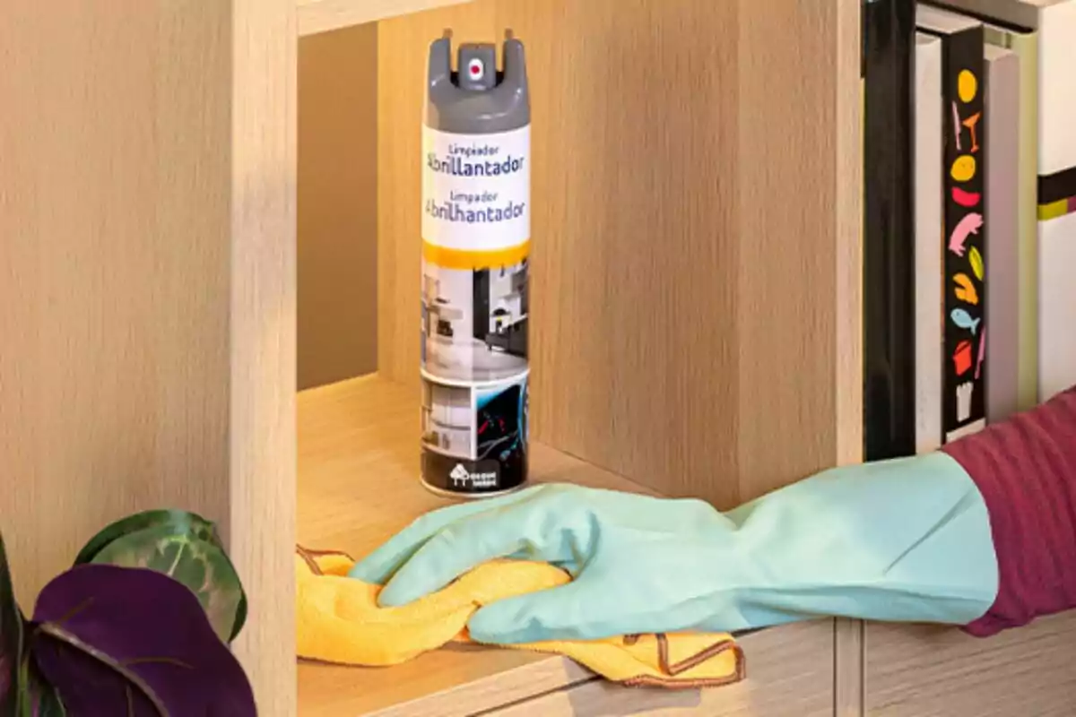 Persona con guante de limpieza azul limpiando un mueble de madera con un trapo amarillo y un aerosol limpiador abrillantador sobre el estante Persona con guante de limpieza azul limpiando un mueble de madera con un trapo amarillo y un aerosol limpiador abrillantador sobre el estante