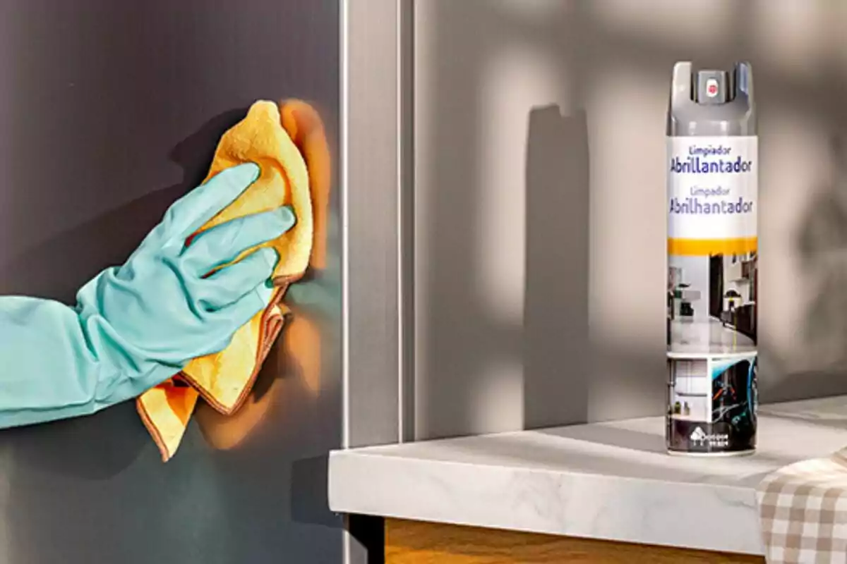 Persona con guante azul limpiando una superficie de acero inoxidable con un paño amarillo y un aerosol limpiador sobre una encimera blanca Persona con guante azul limpiando una superficie de acero inoxidable con un paño amarillo y un aerosol limpiador sobre una encimera blanca