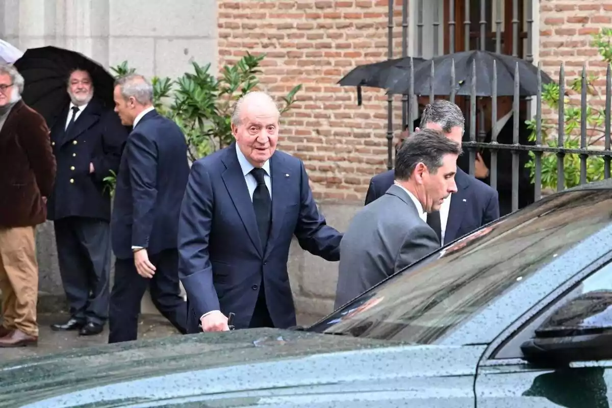 Un grupo de hombres, algunos con paraguas, se encuentran en la calle junto a un coche mientras uno de ellos, vestido con traje oscuro, se dispone a entrar en el vehículo.