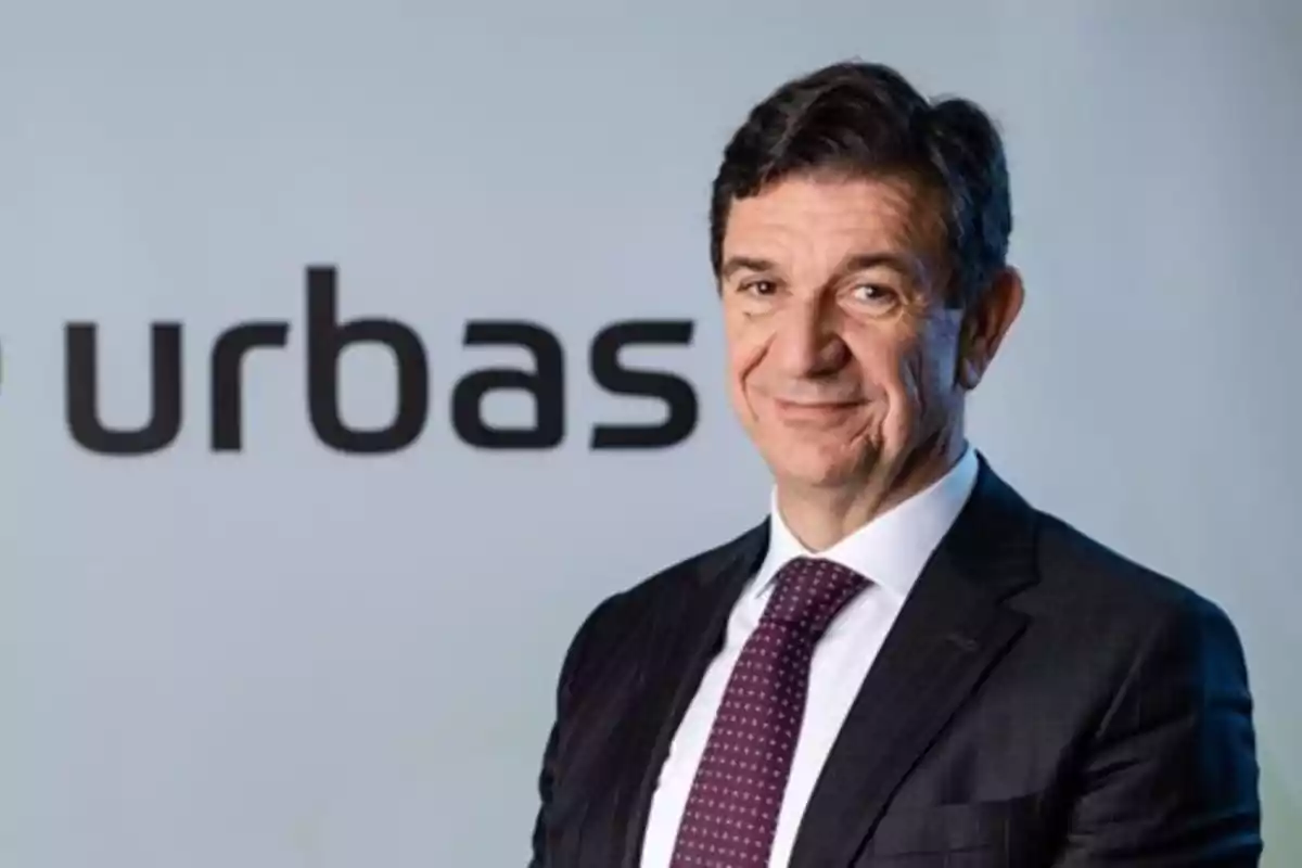 Un hombre con traje y corbata sonríe frente a un fondo con el logotipo de "urbas".