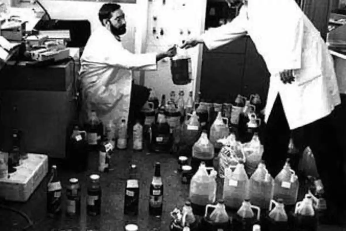 Dos personas con batas de laboratorio están en una habitación llena de botellas y recipientes, una de ellas está sentada y la otra de pie, mientras intercambian un objeto.