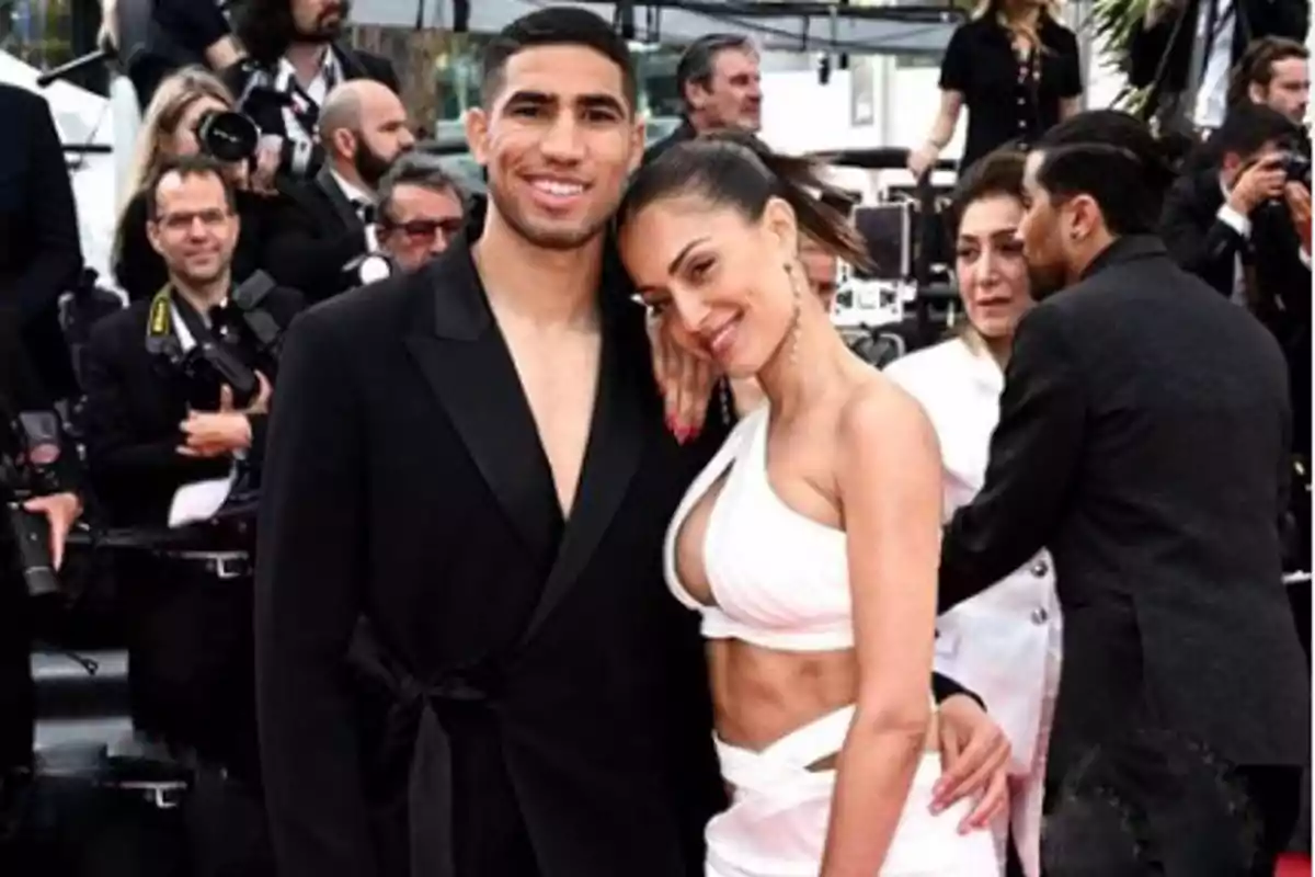 Una pareja posando para una foto en un evento con varias personas y fotógrafos en el fondo.