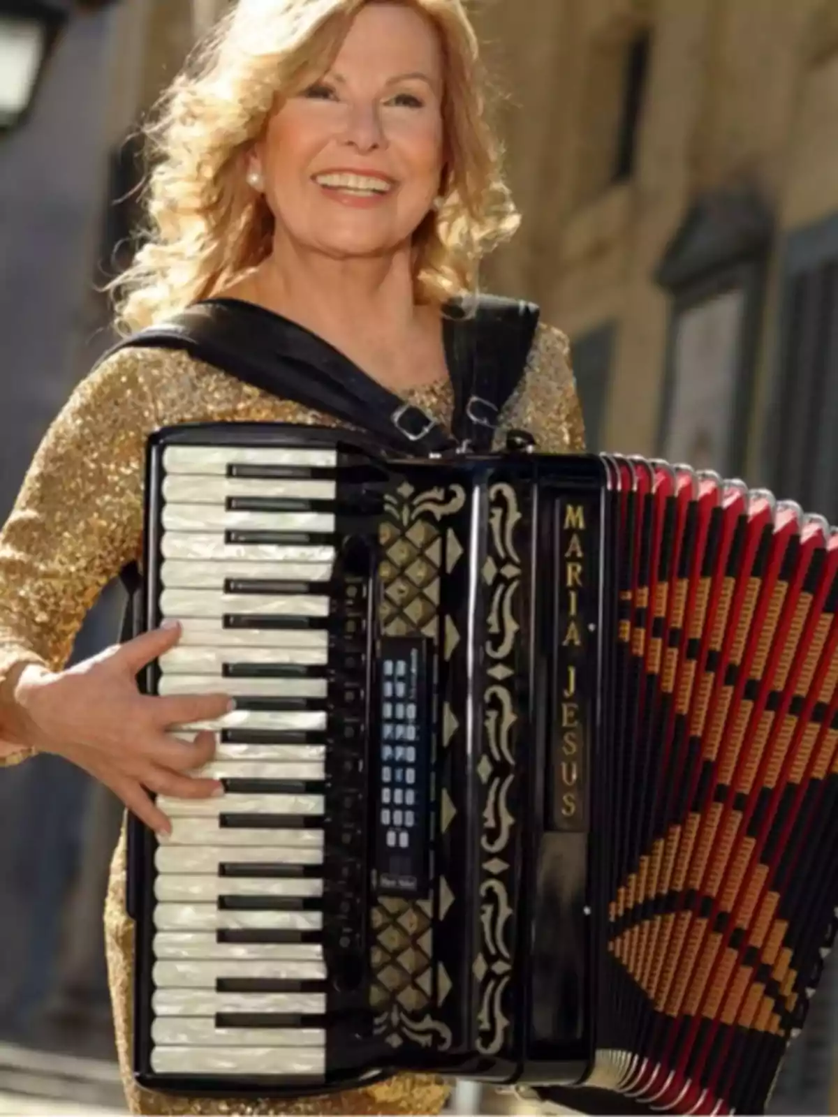 Una mujer sonriente con cabello rubio y rizado, vestida con un traje dorado brillante, toca un acordeón negro con detalles dorados y rojos.