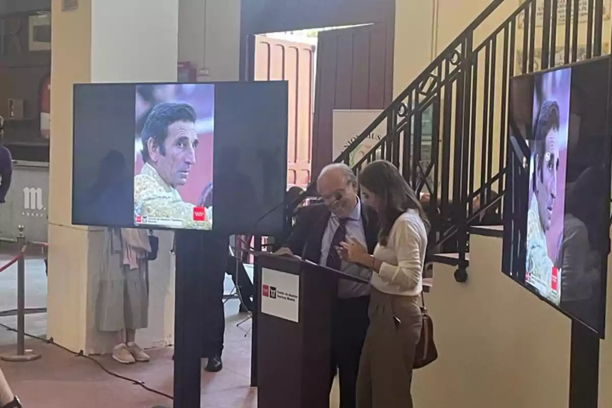 Dos personas están de pie junto a un podio, con dos pantallas grandes que muestran la imagen de un hombre en traje de luces.