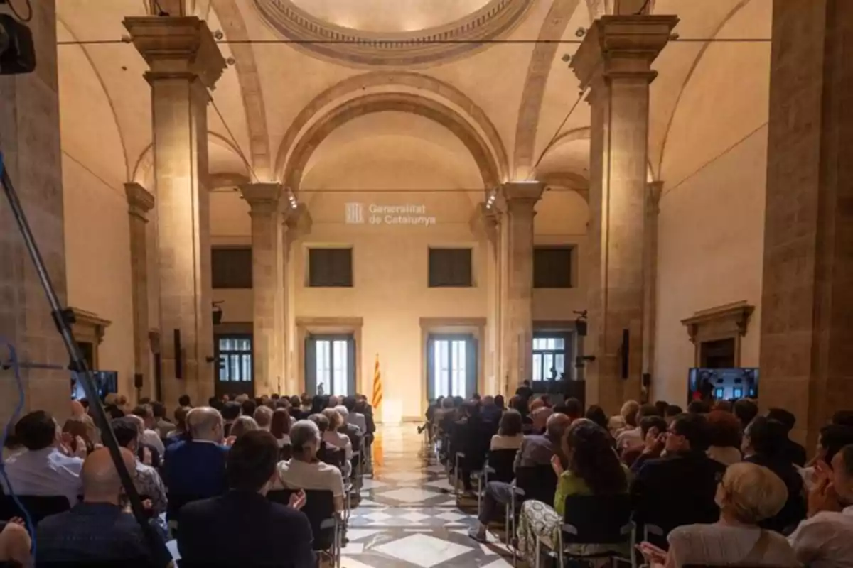 Una audiencia sentada en un salón con columnas y una bandera catalana al fondo.