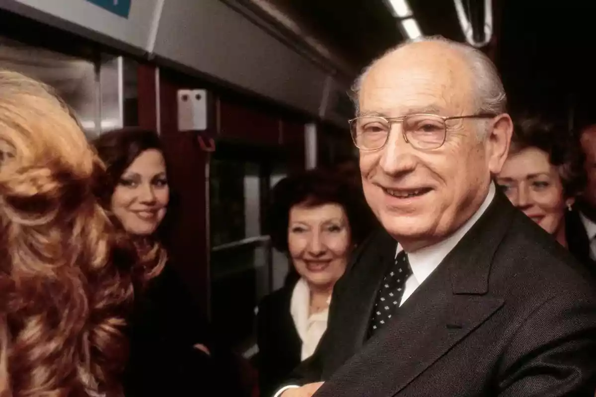 Un hombre mayor con gafas y traje sonríe mientras está rodeado de varias personas en un tren.