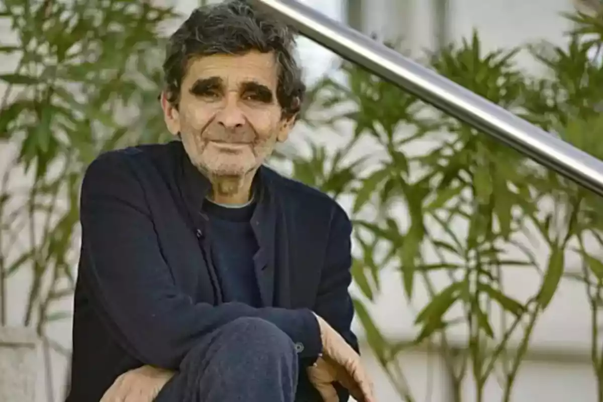 Un hombre de cabello oscuro y barba ligera, sentado al aire libre con plantas de fondo.