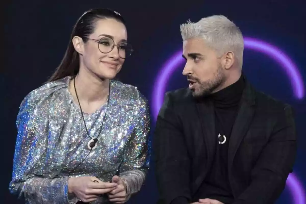 Adara Molinero y Miguel Frigenti sentados en el plató de 'GH' con rostros sonrientes hablando entre ellos