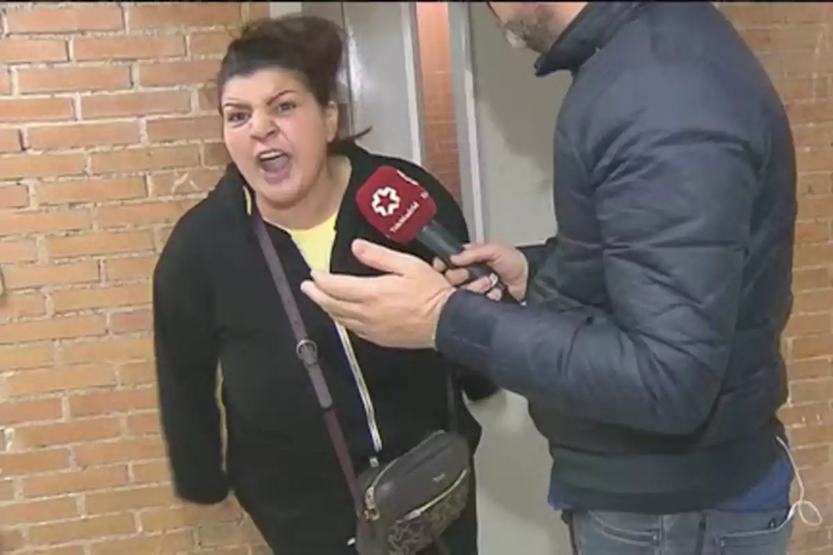 Mujer con expresión molesta siendo entrevistada por un reportero con micrófono rojo frente a una pared de ladrillos Mujer con expresión molesta siendo entrevistada por un reportero con micrófono rojo frente a una pared de ladrillos