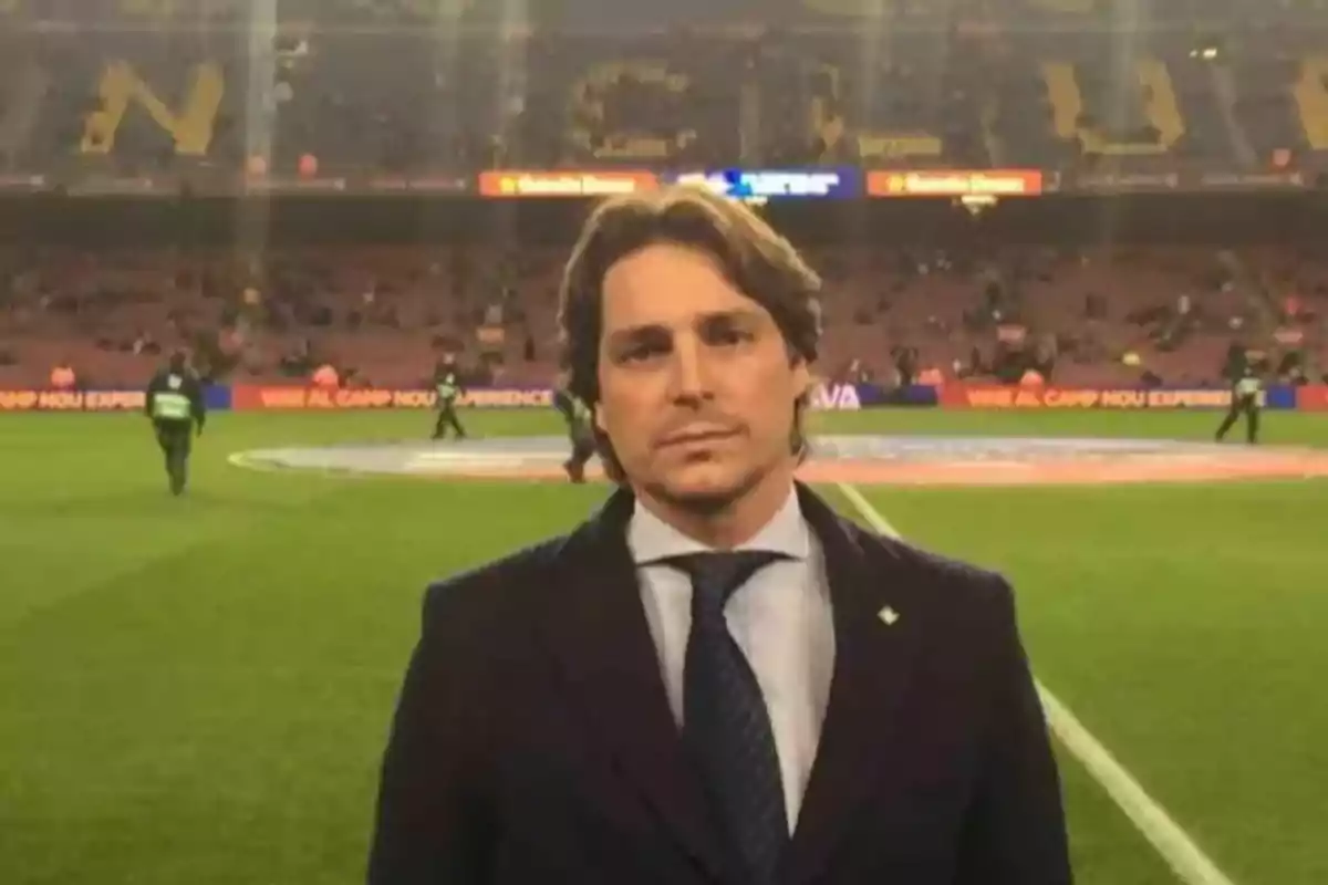 Un hombre con traje y corbata está de pie en un estadio de fútbol, con el campo y las gradas de fondo.