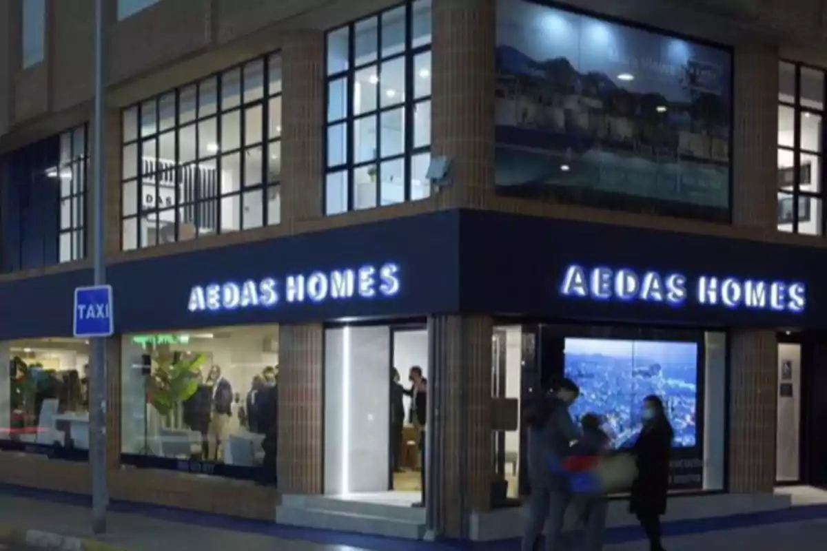 Fachada de una oficina de AEDAS Homes iluminada por la noche con personas caminando frente a la entrada y un letrero de taxi en la acera Fachada de una oficina de AEDAS Homes iluminada por la noche con personas caminando frente a la entrada y un letrero de taxi en la acera