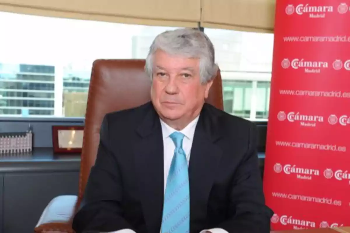 Hombre de cabello canoso con traje oscuro y corbata celeste sentado en una oficina junto a un cartel rojo de la Cámara de Madrid