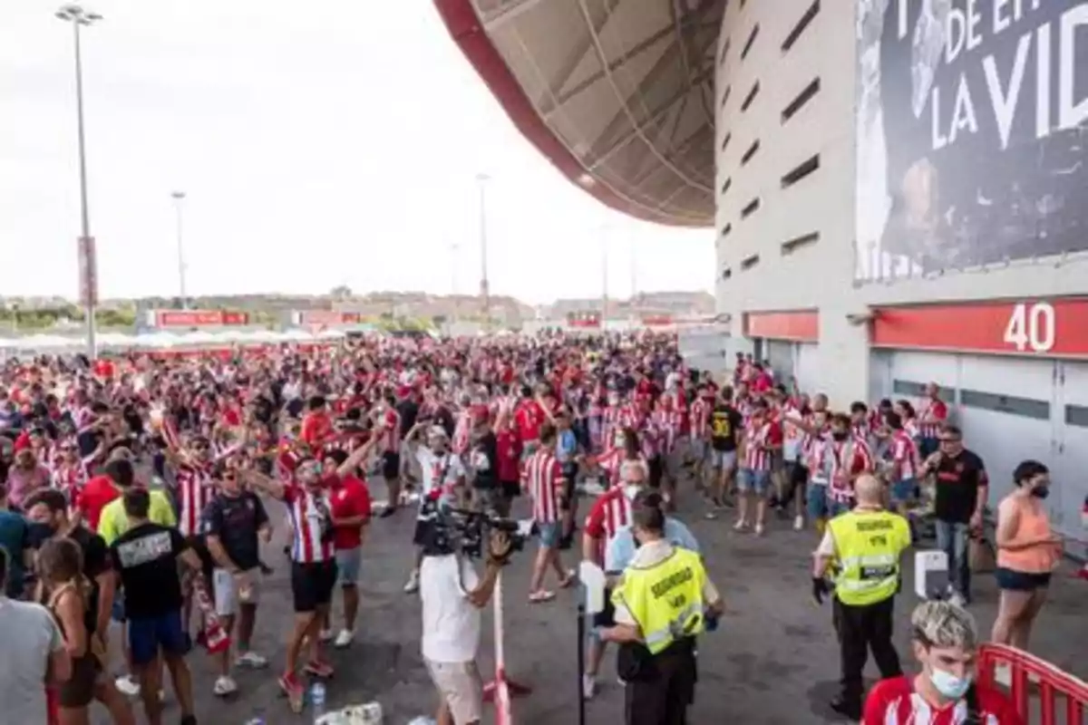 Aficionados con camisetas a rayas rojas y blancas se congregan fuera de un estadio de fútbol, con personal de seguridad presente.