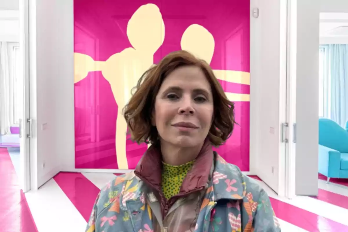 Una mujer con cabello corto y castaño está de pie frente a una pared con un diseño artístico en tonos rosa y beige, vistiendo una chaqueta colorida con patrones florales y geométricos.