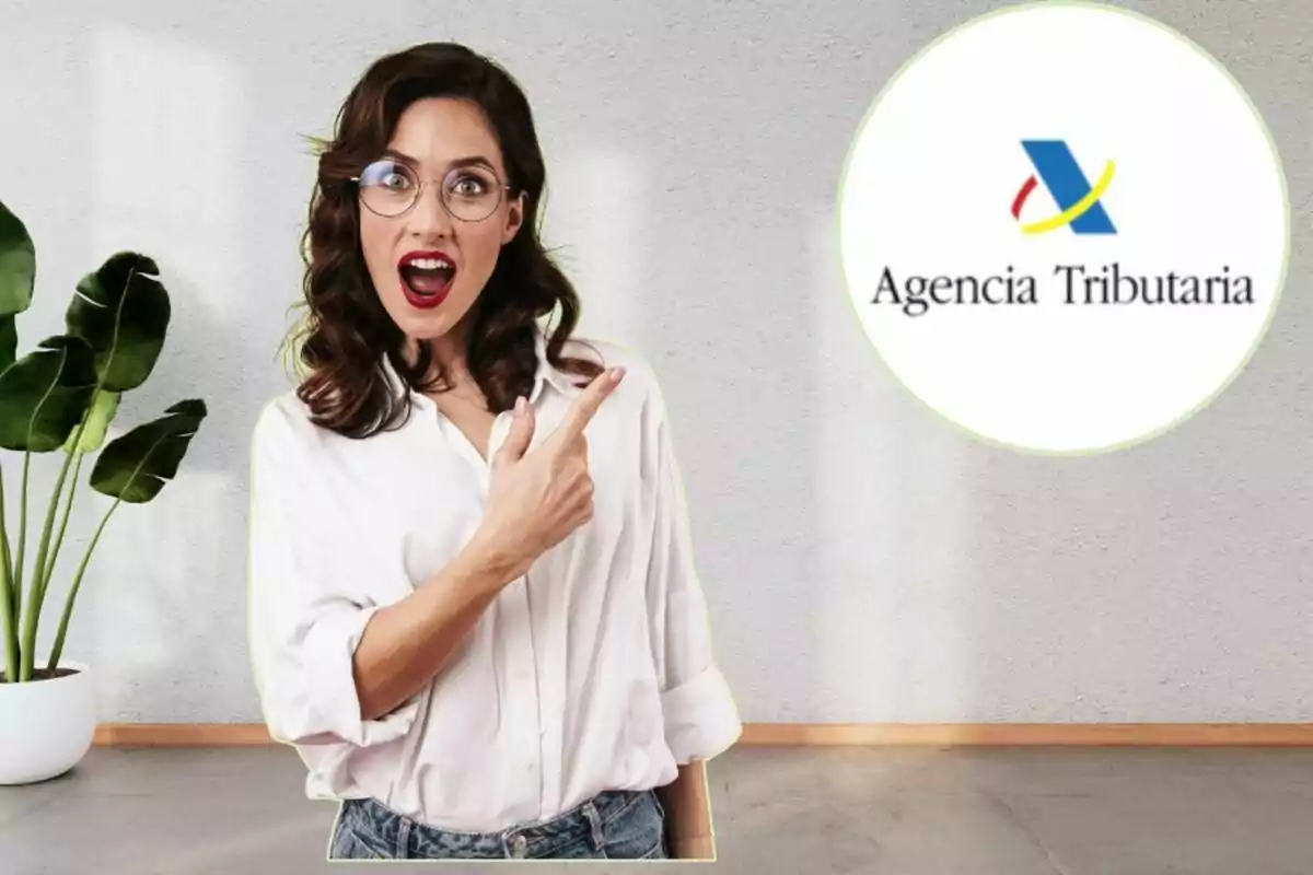 Mujer sorprendida con gafas y camisa blanca señalando el logo de la Agencia Tributaria en un fondo interior con planta.