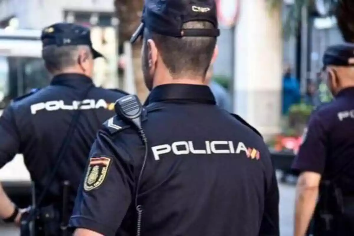 Tres policías con uniformes negros y la palabra "POLICÍA" en la espalda patrullan una calle.