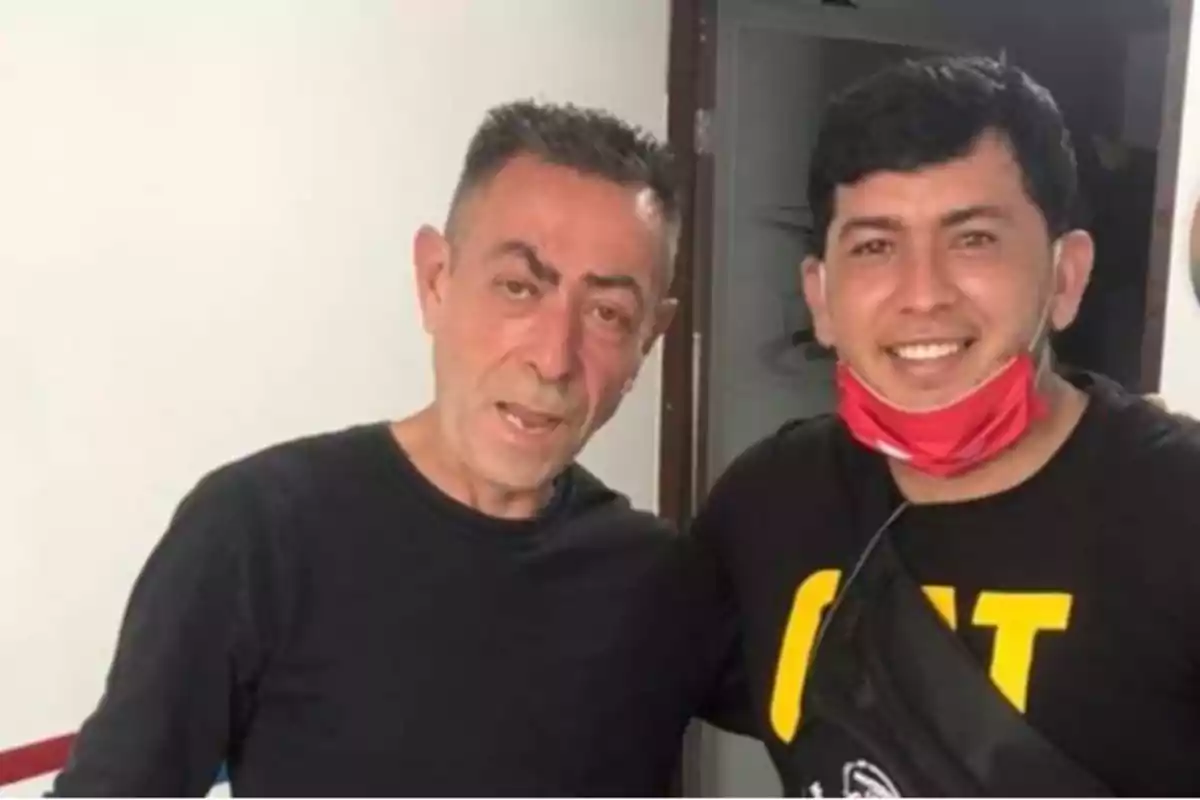 Dos hombres posando juntos, uno de ellos lleva una mascarilla roja bajada al cuello.