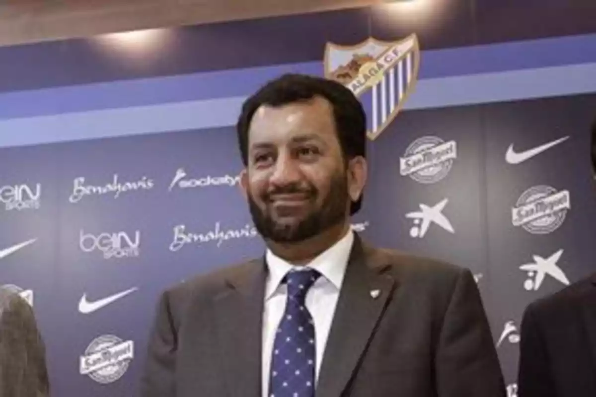 Un hombre con traje y corbata sonríe frente a un fondo con logotipos de marcas y el escudo del Málaga CF.