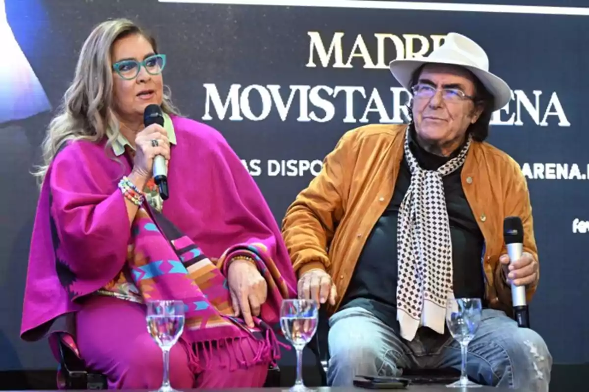 Dos personas están sentadas en un escenario, una de ellas sostiene un micrófono y lleva un atuendo colorido, mientras que la otra persona lleva un sombrero blanco y una chaqueta marrón.