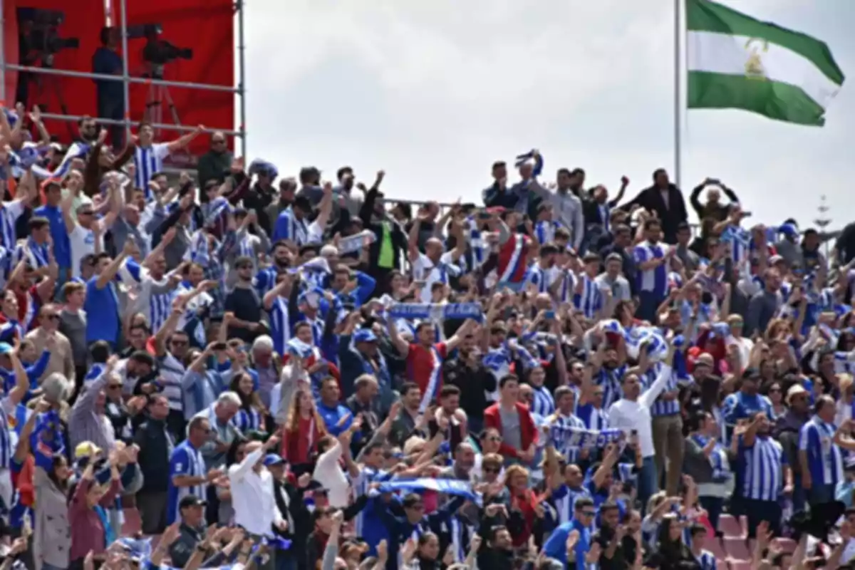 Aficionados animando en un estadio con camisetas a rayas azules y blancas y una bandera verde y blanca ondeando al fondo. Aficionados animando en un estadio con camisetas a rayas azules y blancas y una bandera verde y blanca ondeando al fondo.