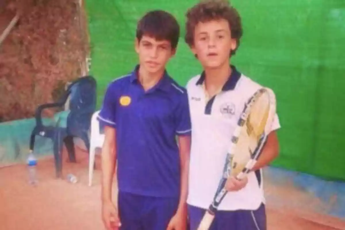 Dos niños en una cancha de tenis, uno de ellos sostiene una raqueta.
