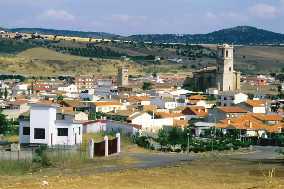Vista panorámica de un pueblo con casas de techos rojos, una iglesia y colinas en el fondo. Vista panorámica de un pueblo con casas de techos rojos, una iglesia y colinas en el fondo.