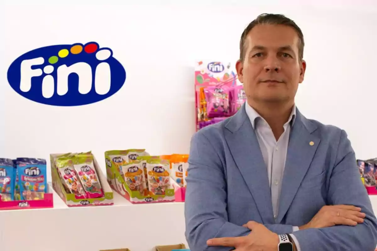 Hombre de traje azul posando frente a un estante con productos de la marca Fini y el logo de la empresa en la pared.