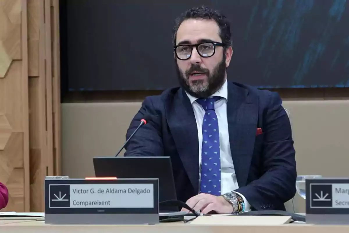 Un hombre con barba y gafas está sentado en una mesa con un micrófono frente a él, vistiendo un traje oscuro y corbata azul con un patrón, en un entorno formal.