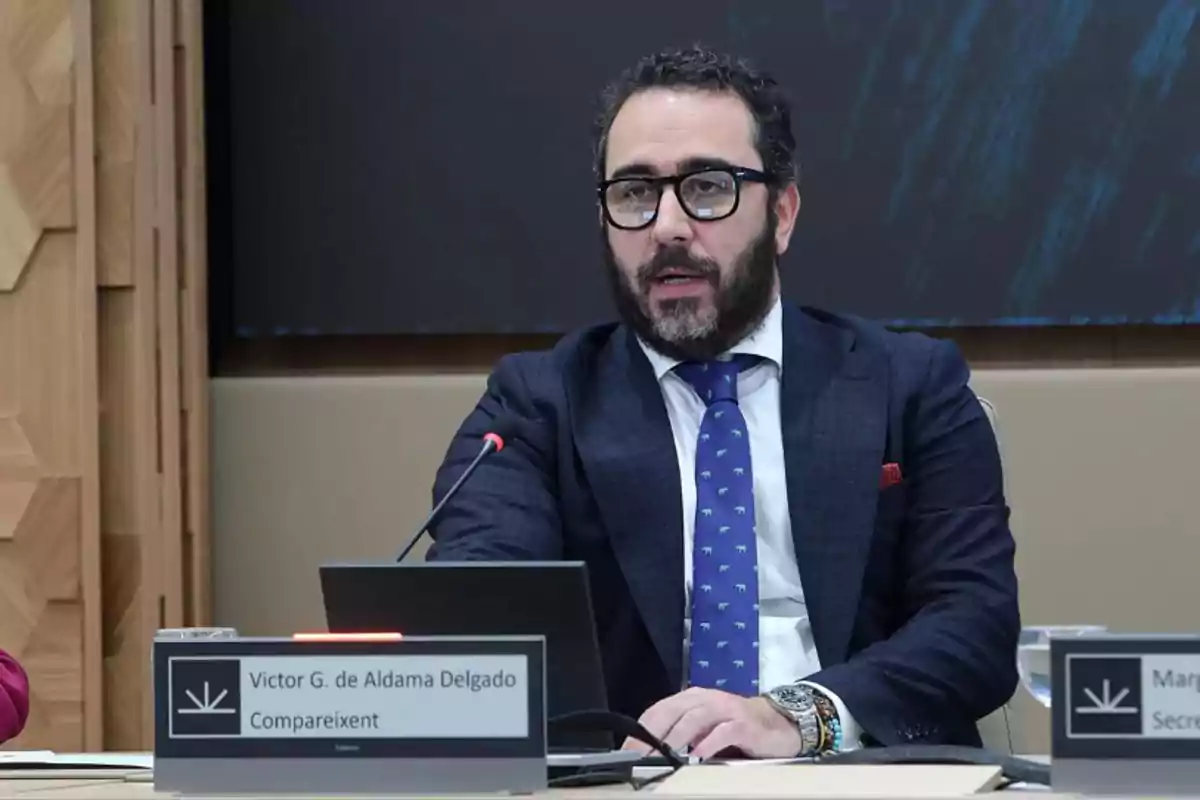 Un hombre con barba y gafas está sentado en una mesa con un micrófono frente a él, vistiendo un traje oscuro y corbata azul con un patrón, en un entorno formal.