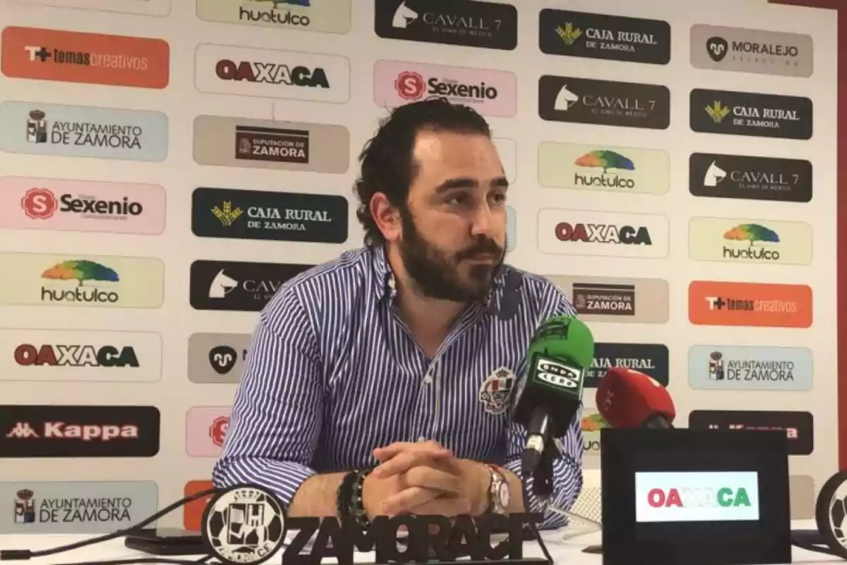 Un hombre con barba y camisa a rayas habla en una conferencia de prensa con micrófonos frente a él y un fondo con logotipos de patrocinadores.