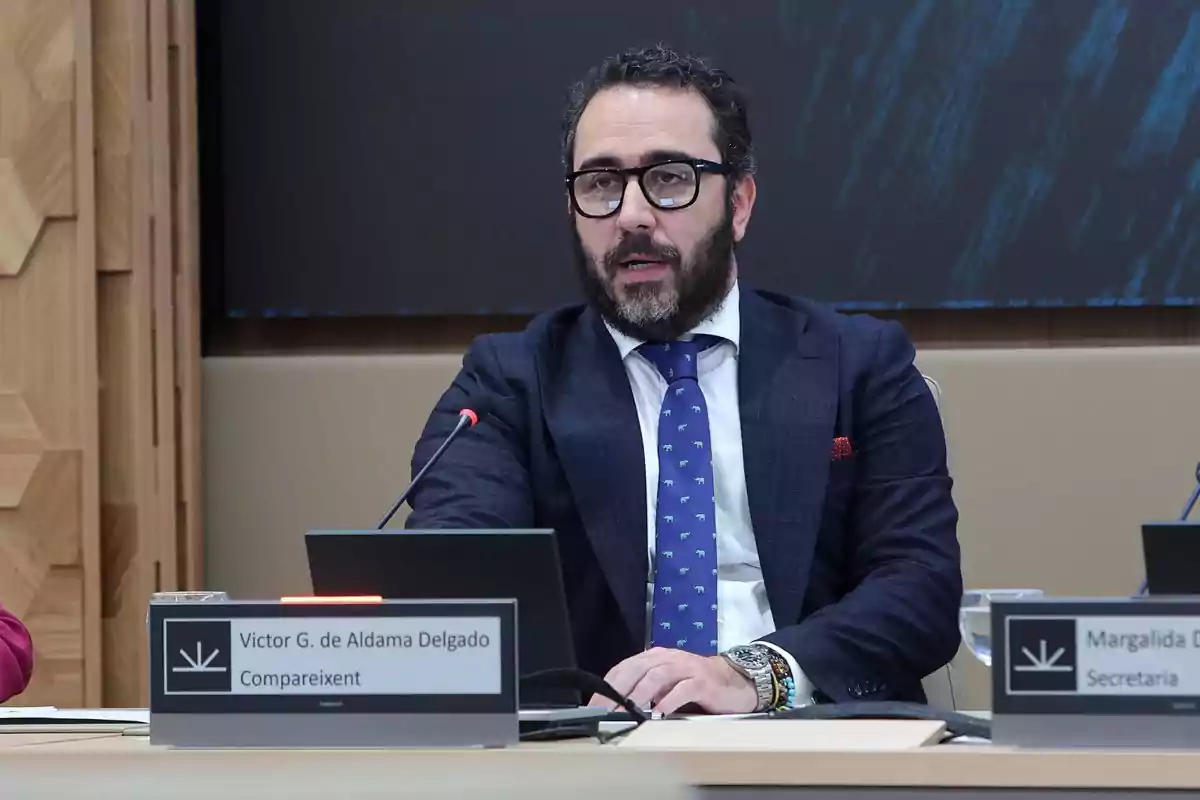 Un hombre con barba y gafas está sentado en una mesa con un micrófono frente a él, vistiendo un traje oscuro y corbata azul con un diseño de elefantes.