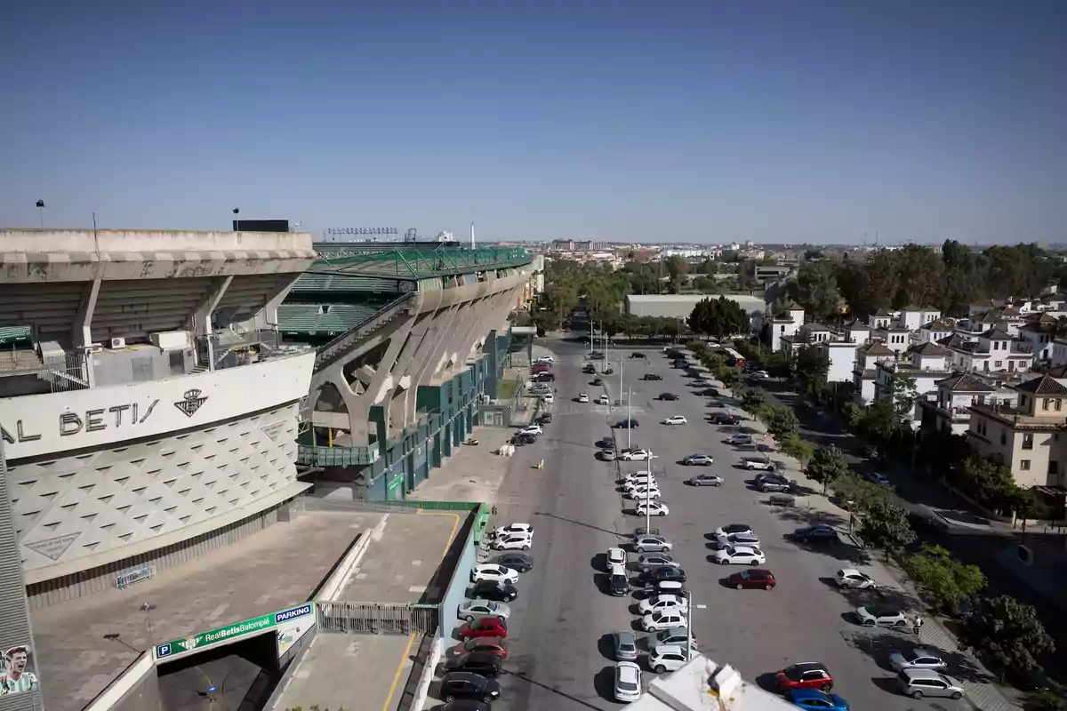 Vista aérea de un estadio de fútbol con un aparcamiento y una zona residencial al fondo en un día soleado.