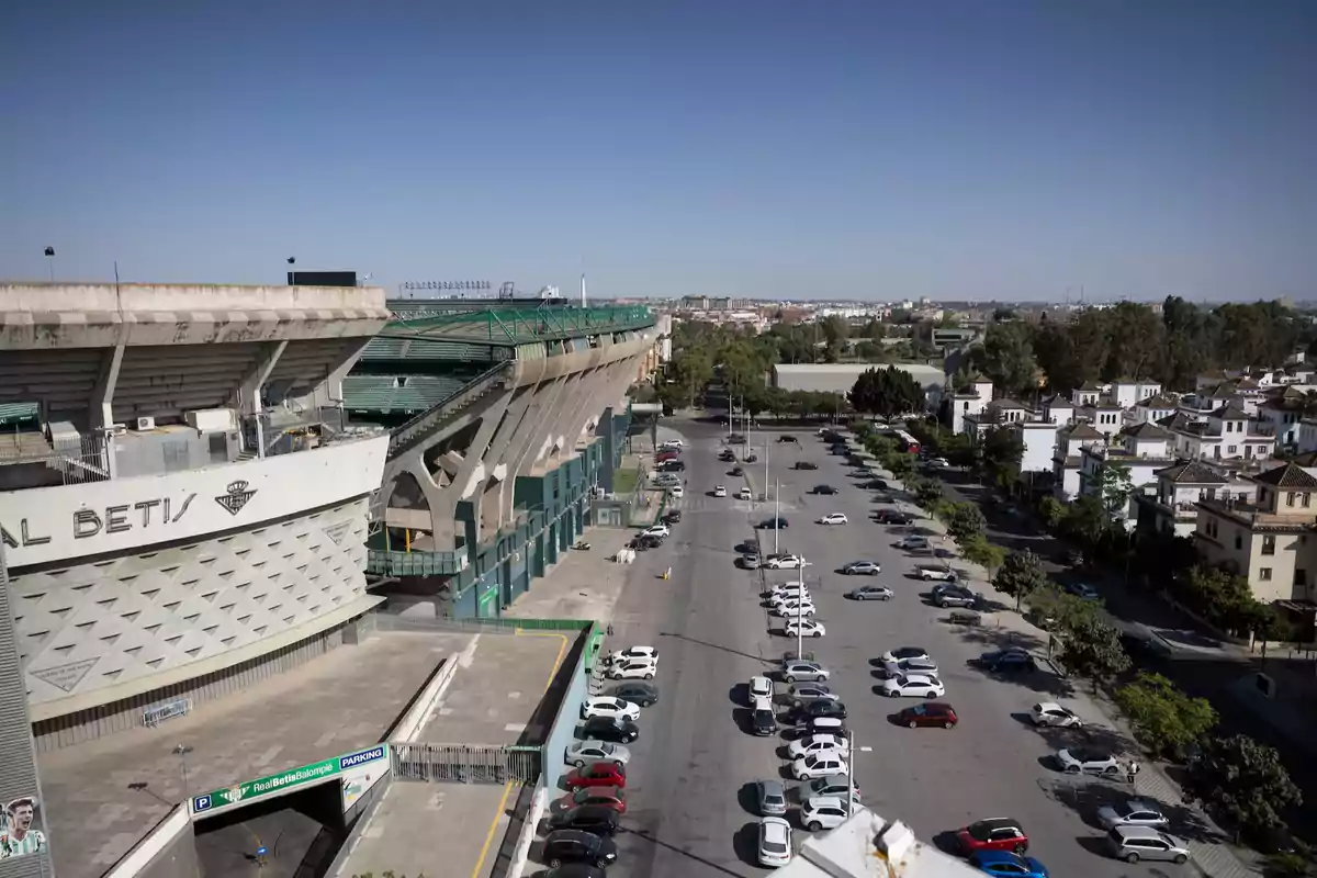 Vista aérea de un estadio de fútbol con un aparcamiento lleno de coches y una zona residencial al fondo.